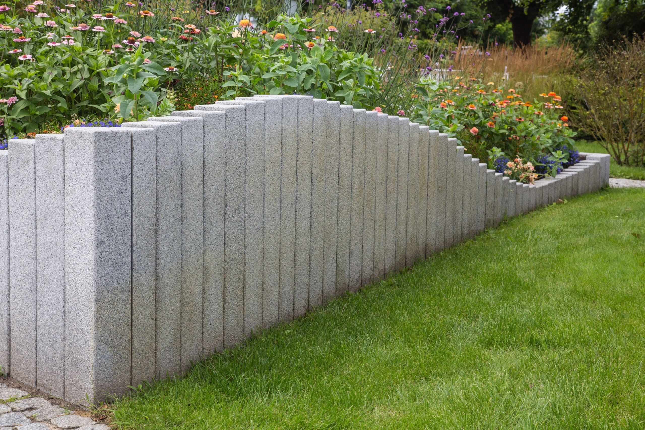 Muret d'escalier en granite gris texturé délimitant une pelouse verte et un parterre de zinnias roses et oranges.