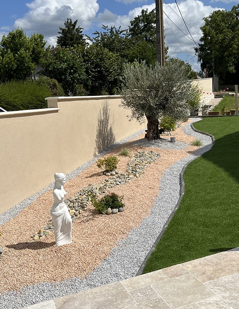 Aménagement paysager méditerranéen avec statue blanche, gravier ocre et gris, olivier, et gazon synthétique près d'un mur beige.