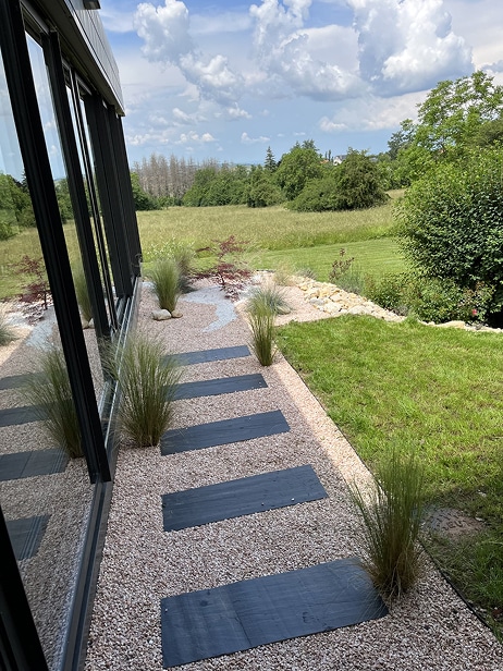 Allée moderne bordant une façade vitrée, avec des pas japonais noirs sur gravier clair et des touffes d'herbes ornementales, vue sur un paysage verdoyant.