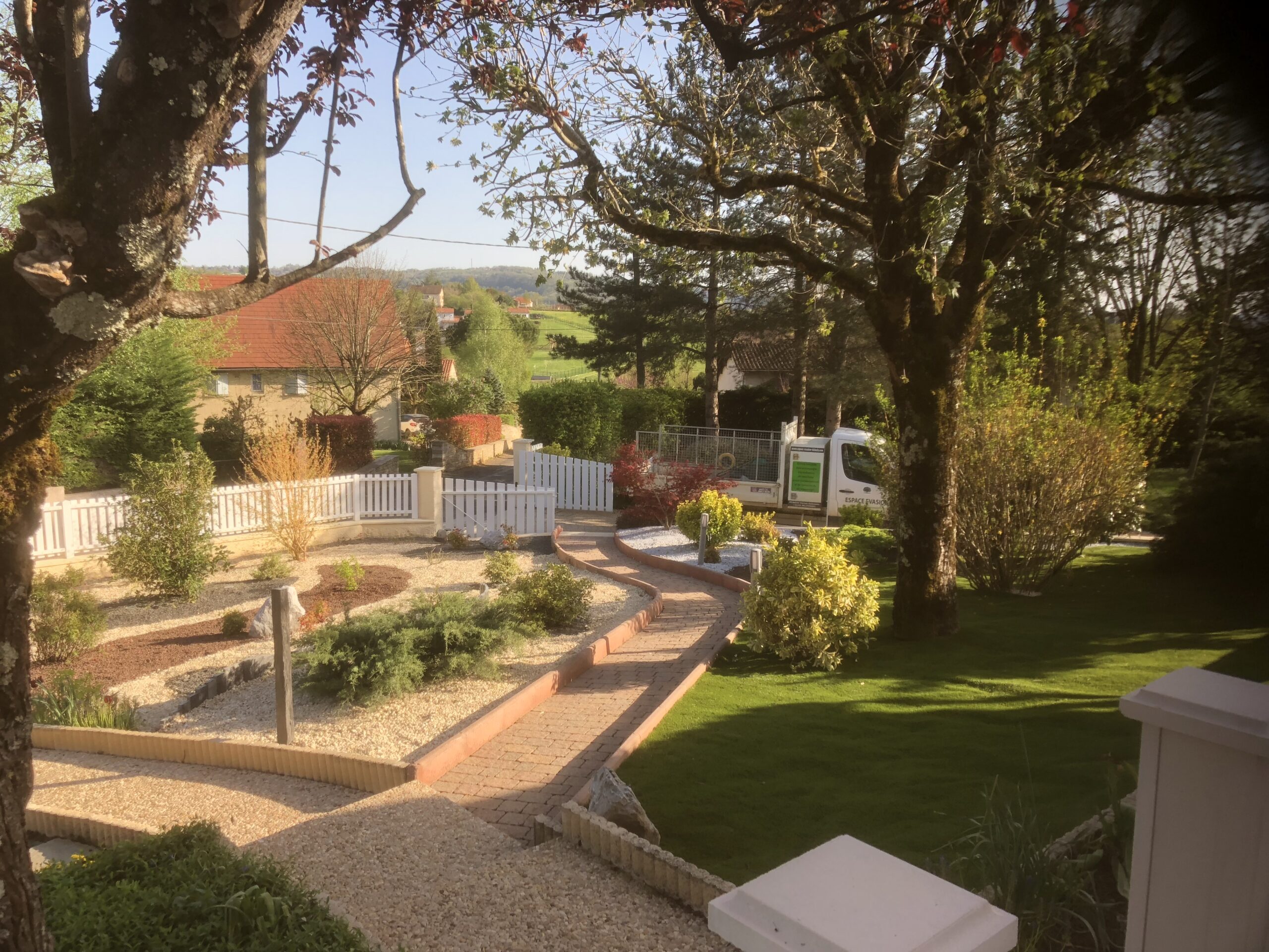 Jardin sec avec olivier, statue et gravier coloré Aménagement paysager méditerranéen avec statue blanche, gravier ocre et gris, olivier, et gazon synthétique près d'un mur beige.