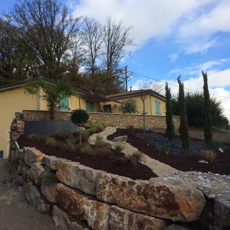 Jardin sec avec olivier, statue et gravier coloré Aménagement paysager méditerranéen avec statue blanche, gravier ocre et gris, olivier, et gazon synthétique près d'un mur beige.