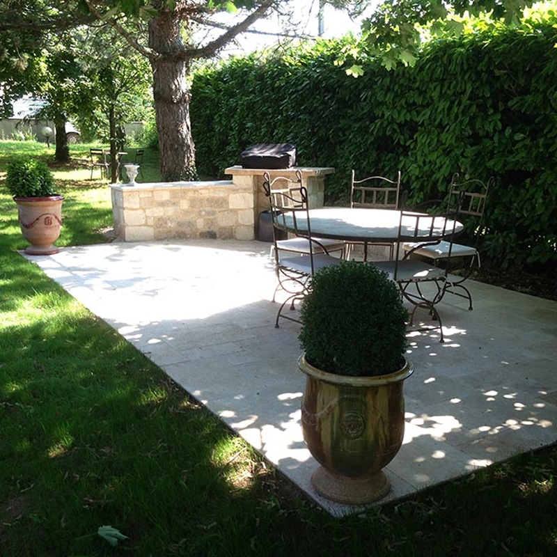 Jardin sec avec olivier, statue et gravier coloré Aménagement paysager méditerranéen avec statue blanche, gravier ocre et gris, olivier, et gazon synthétique près d'un mur beige.