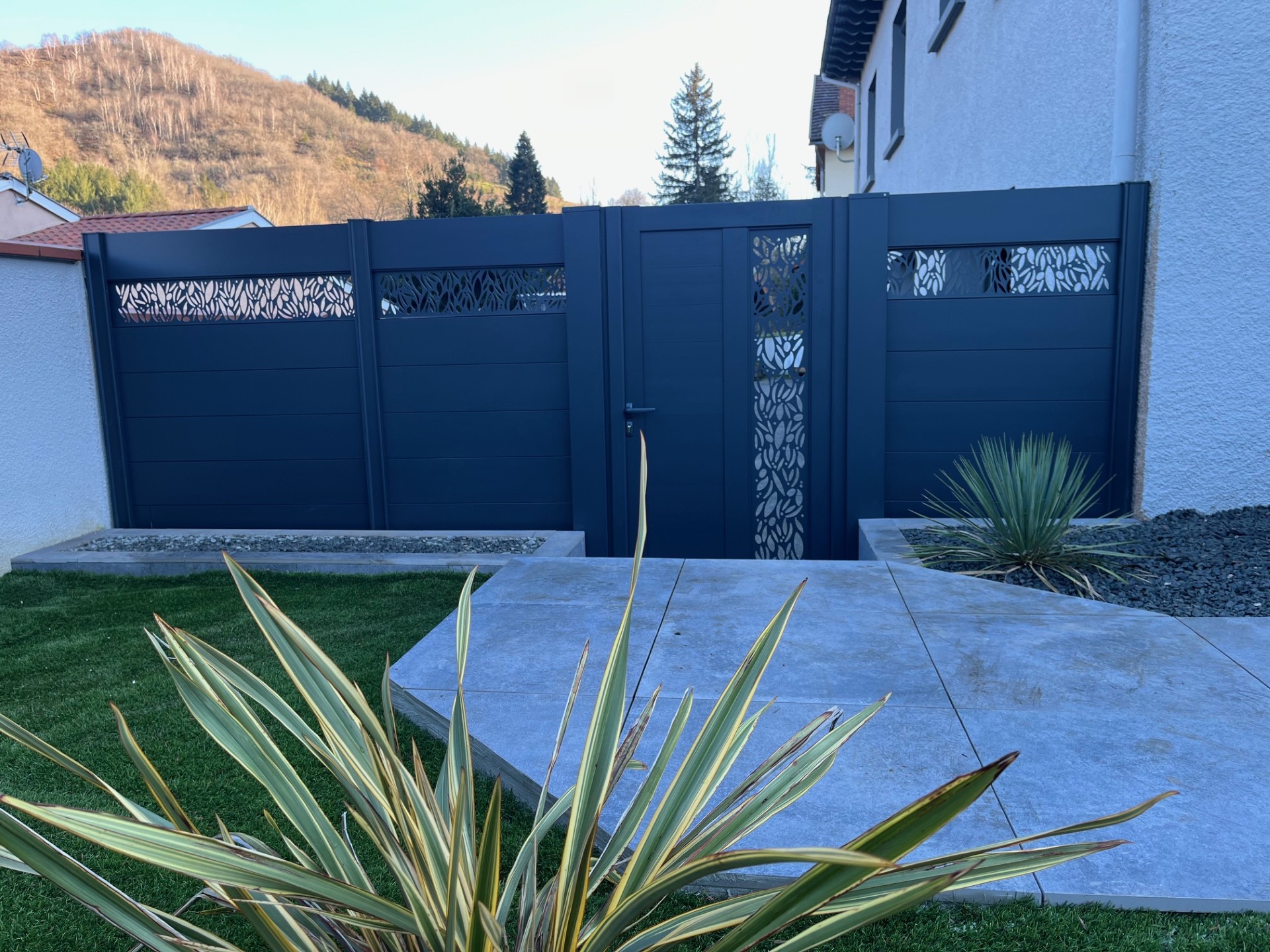 Jardin sec avec olivier, statue et gravier coloré Aménagement paysager méditerranéen avec statue blanche, gravier ocre et gris, olivier, et gazon synthétique près d'un mur beige.