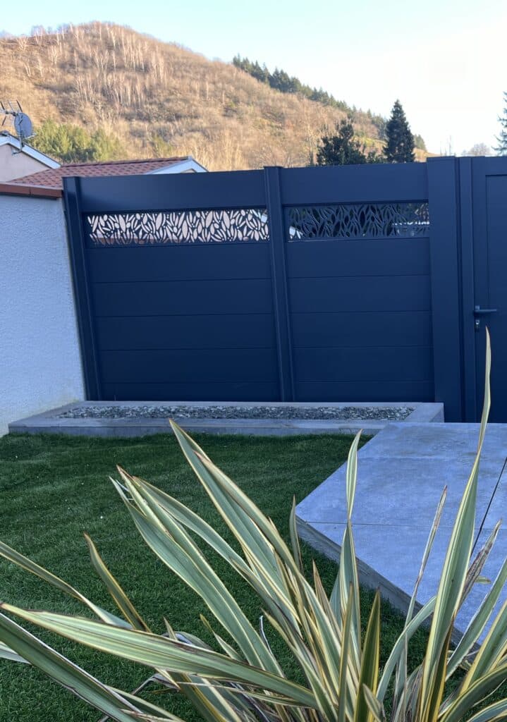 Portail Bleu Nuit Moderne avec Découpe Laser Design Portail ajouré bleu nuit moderne avec motifs végétaux stylisés, devant une pelouse et une plante panachée, sur fond de colline boisée.