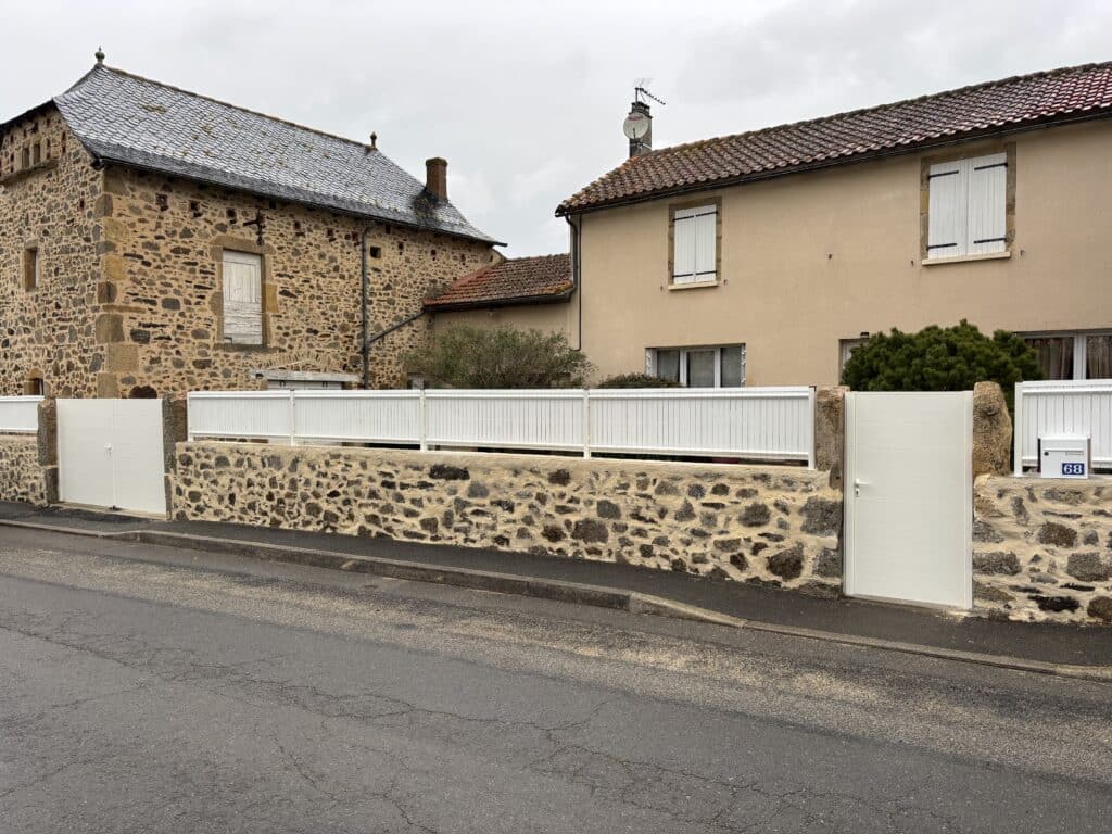 Portail blanc, maison pierre et crépie, clôture neuve Façades de deux maisons, l'une en pierre et l'autre crépie, séparées par une clôture blanche neuve et un portail moderne devant un mur de pierres sèches. Numéro de boîte aux lettres 68.